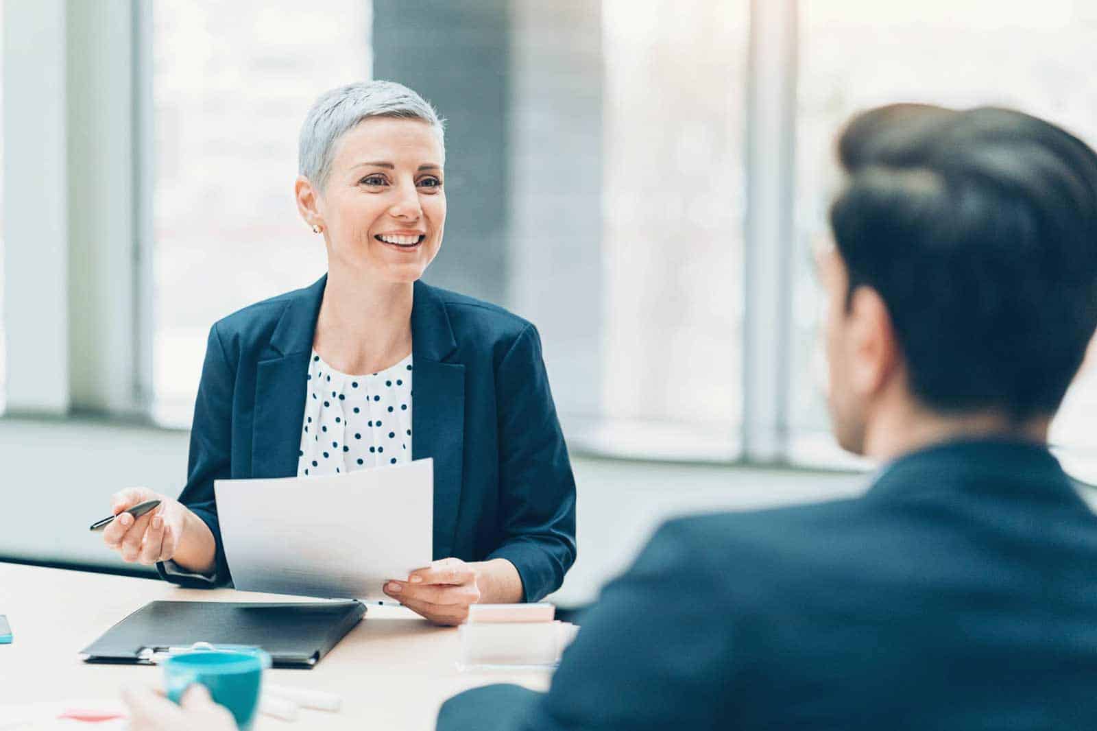 interview guide - a grey haired women and black haired man chatting at a work desk - the woman is holding a piece of paper