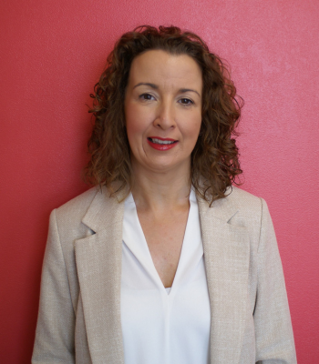 Helen Hardy - female with brown curly hair wearing a white shirt and beige linen blazer standing in front of pink wall